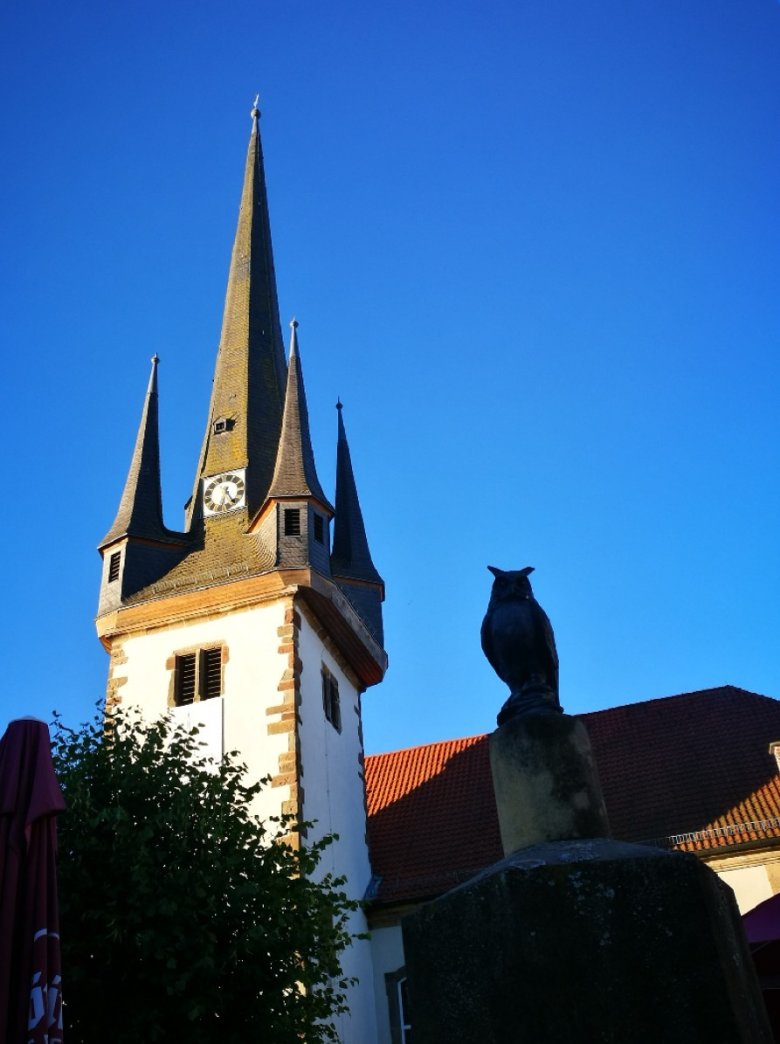 Kirchturm von Oberaula vor blauem Himmel; davor Eule auf dem Marktbrunnen